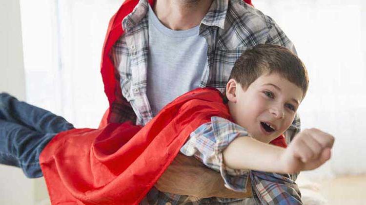 Young boy wearing a cape “flying” in his father’s arms.