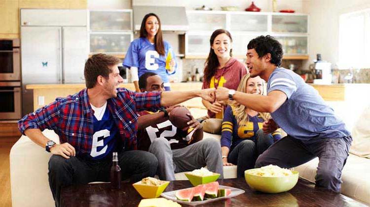 Football fans watching the big game and fist bumping on a couch with snacks.
