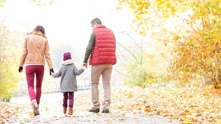 Parents holding their kid's hand walking down a leafy lane in the fall.