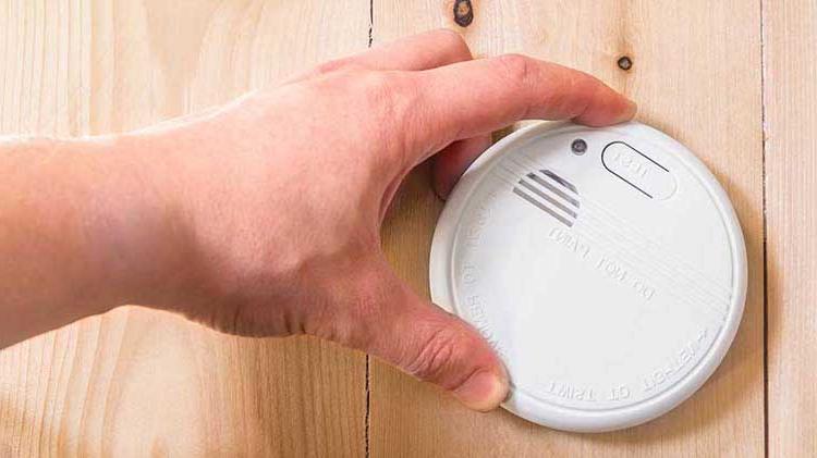 Person's hand holding a smoke detector against a wooden wall.