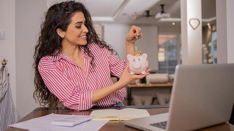 Woman with bills, laptop and calculator in front of her and adding money in a piggy bank.