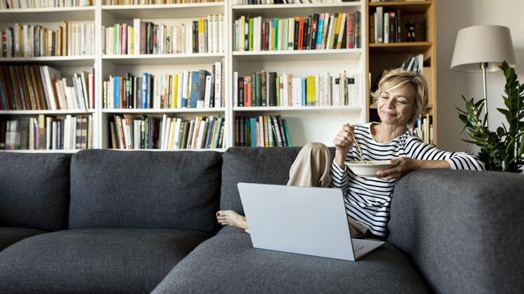 Mature woman using a laptop to work on her digital estate plan and having lunch on a couch.