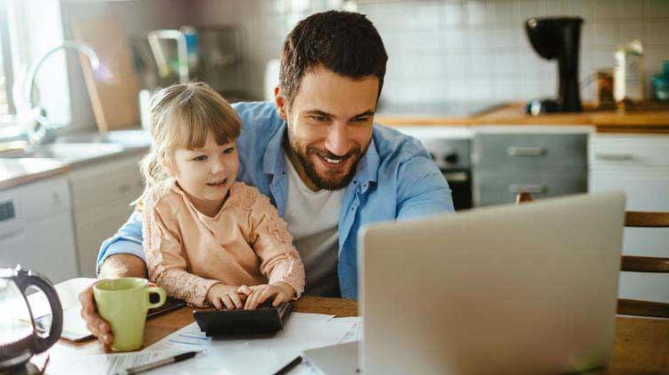A man with his daughter sitting on his lap at the kitchen table working on his laptop exploring different kinds of savings accounts.