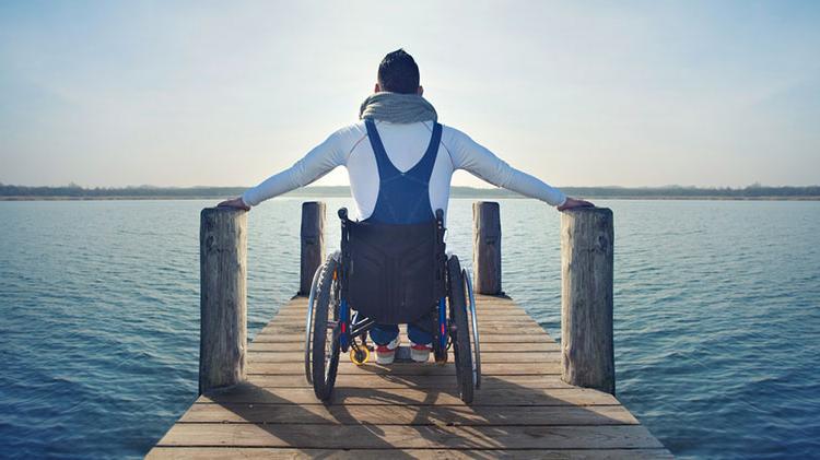 A disabled man sits in his wheelchair on a pier overlooking a lake.