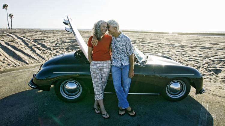 Senior couple leaning on convertible car with a surfboard on the beach.
