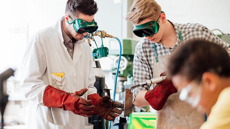 Two young men welding with green safety goggles and red protective gloves.